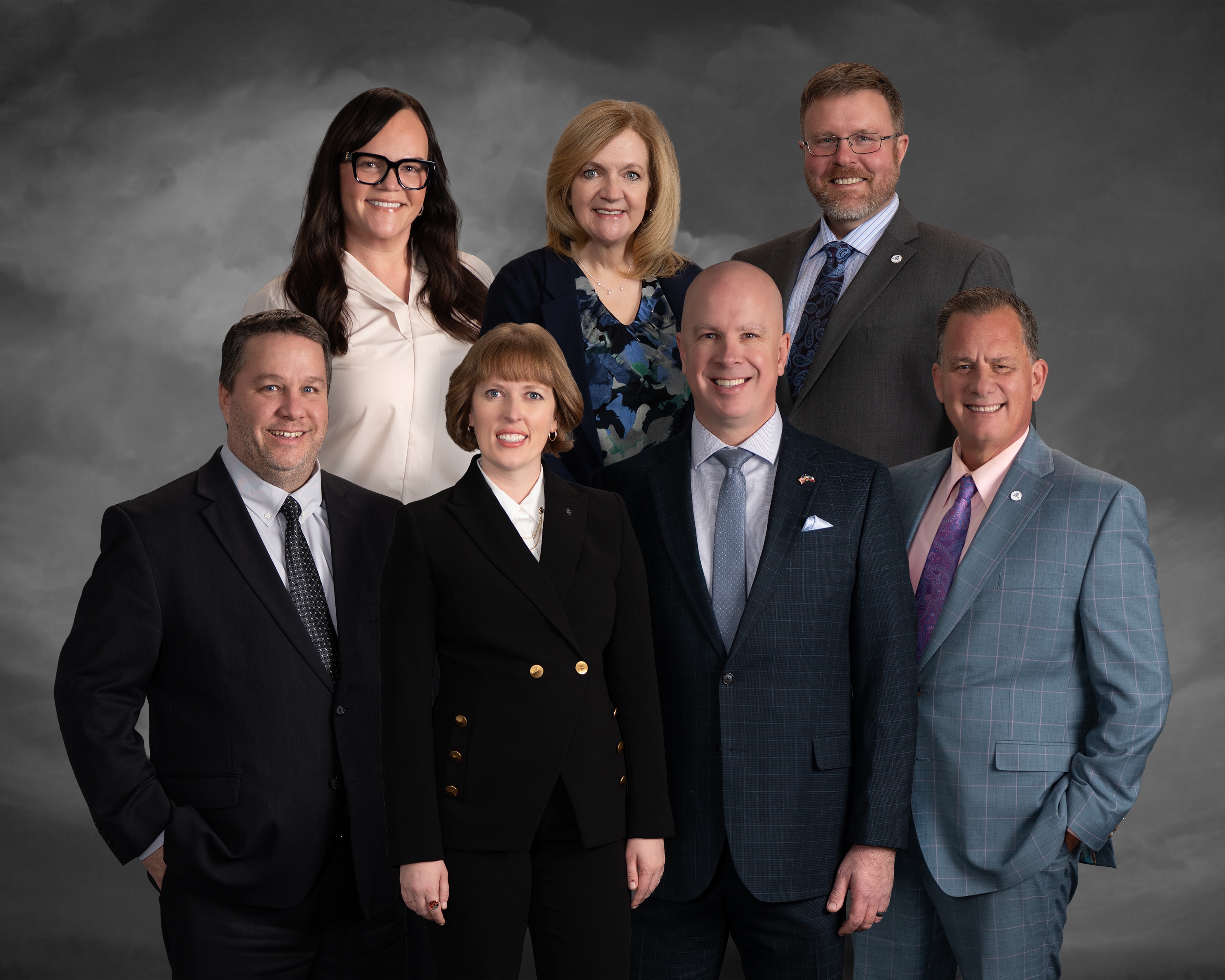 Seven professionally dressed adults, four men and three women, pose together and smile in front of a dark, cloudy studio backdrop.