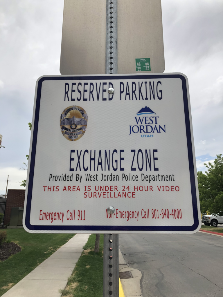 A sign for a reserved parking exchange zone by the West Jordan Police Department. It states the area is under 24-hour video surveillance. Emergency and non-emergency contact numbers are provided.