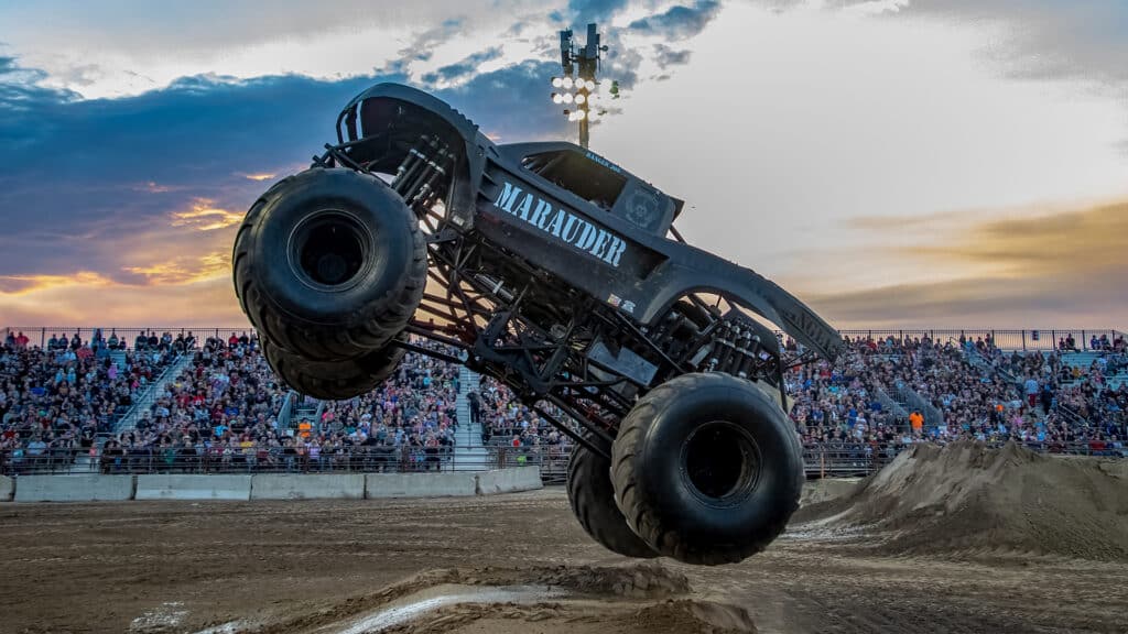 A monster truck labeled Marauder soars through the air during a stunt show, with a large crowd watching from the stands. The sky is a mix of orange and blue hues, indicating a sunset backdrop. The dirt arena shows marks of tire tracks and jumps.