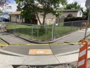 A newly paved concrete sidewalk corner is fenced off with metal barriers and caution tape. A Sidewalk Closed sign is visible. There are two houses in the background with trees and grass.