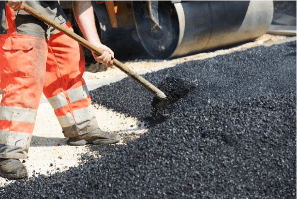 A construction worker in orange pants shovels black asphalt onto a road surface, with a steamroller visible in the background.