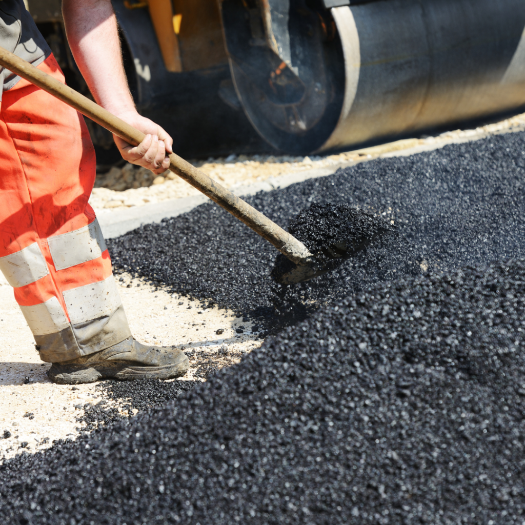 A construction worker wearing orange safety pants spreads fresh asphalt with a shovel on a road surface. A large paving machine is visible in the background.