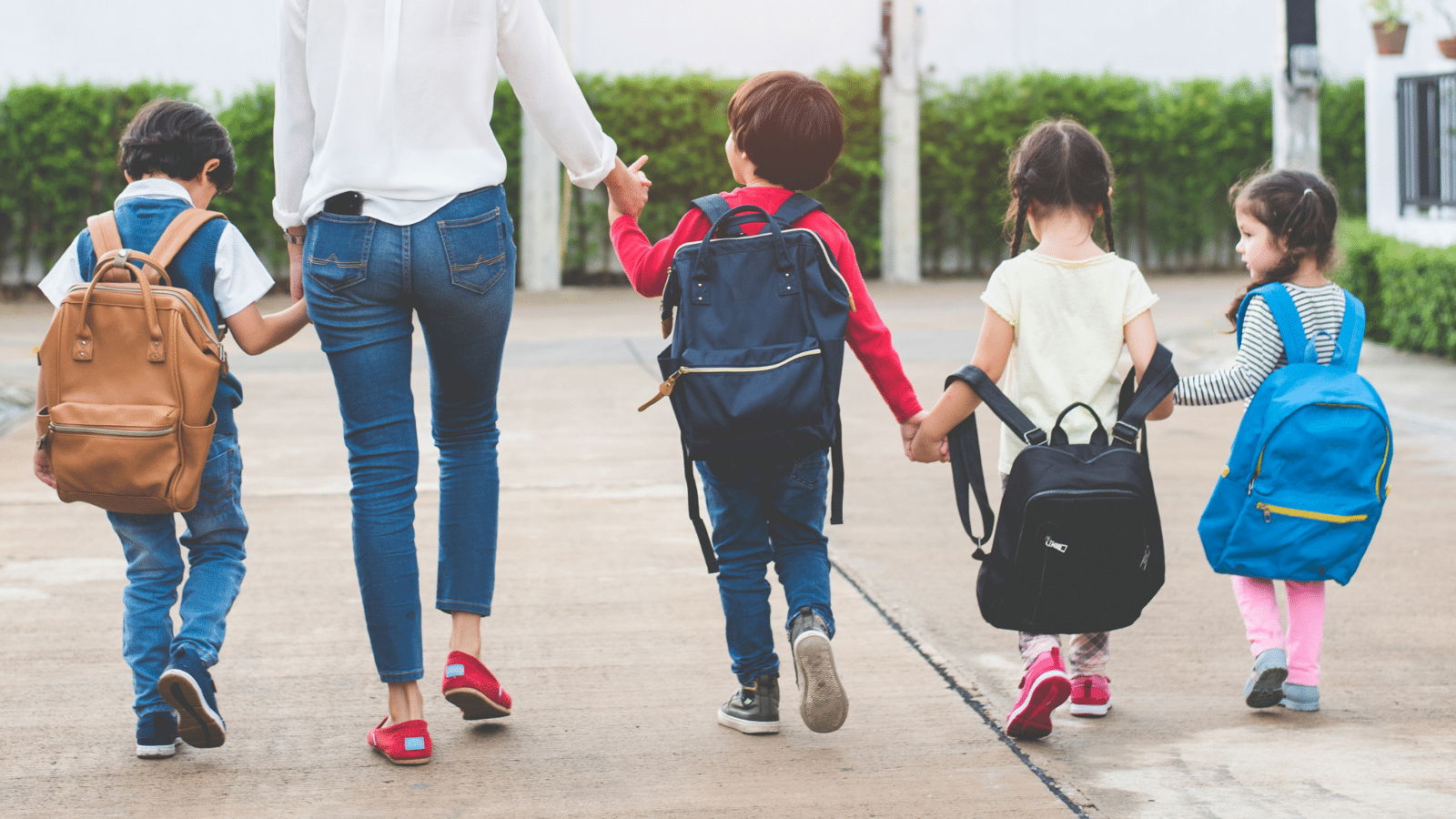 An adult leads four children walking down a path. Each child carries a backpack in different colors: brown, navy blue, black, and light blue. The children hold hands in pairs, and the group walks away from the camera under a clear sky.