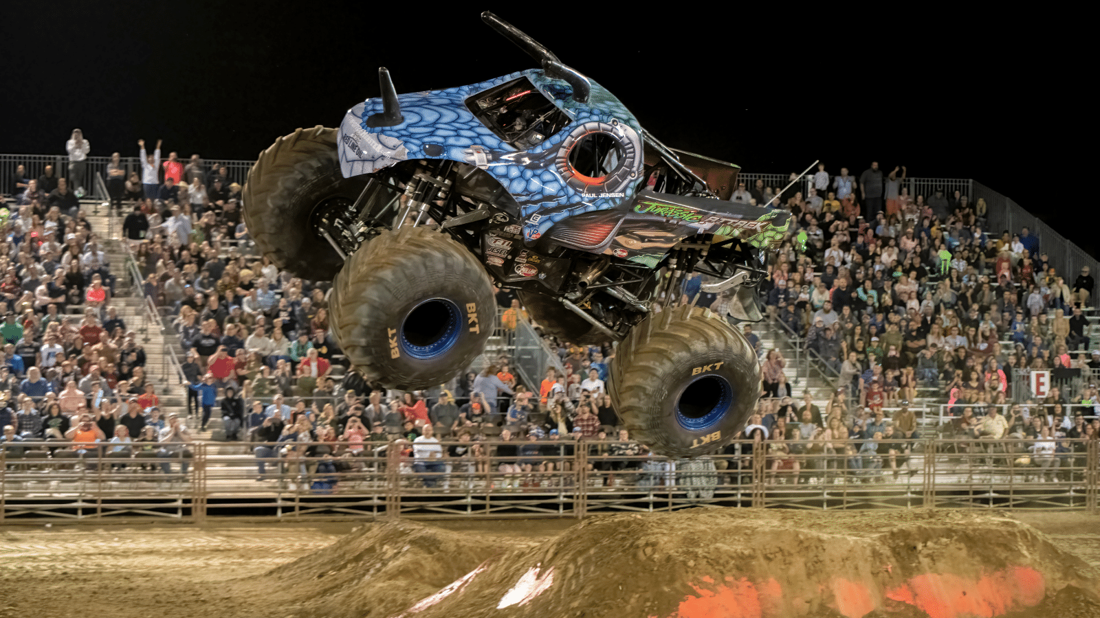 A monster truck with a blue and black design performs a jump over a dirt ramp at an outdoor stadium. The stands are filled with spectators under a night sky.