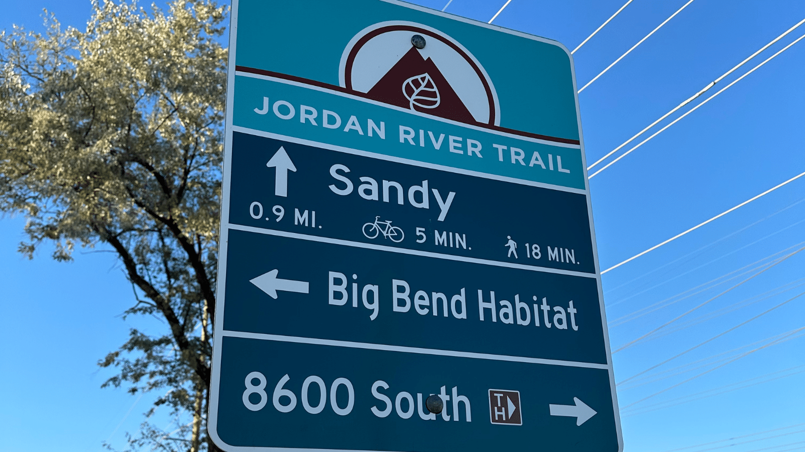 A trail sign showing directions: Jordan River Trail with an arrow to Sandy (0.9 miles, 5 minutes by bike, 18 minutes walking), Big Bend Habitat, and 8600 South. The sky is clear and theres a tree in the background.