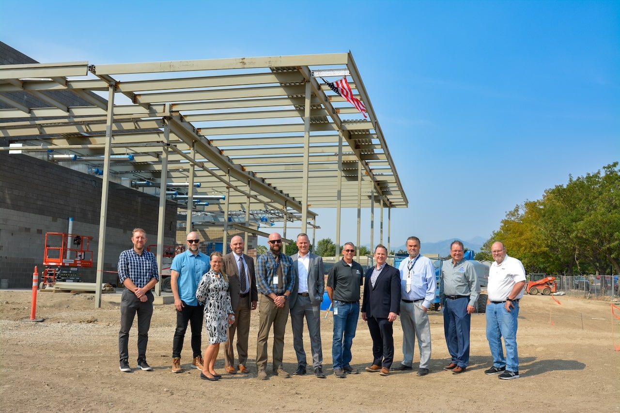 A group of twelve people, including men and women, stand together at a construction site. Theres a steel frame structure in the background, with an American flag hanging from it. The sky is clear and blue.