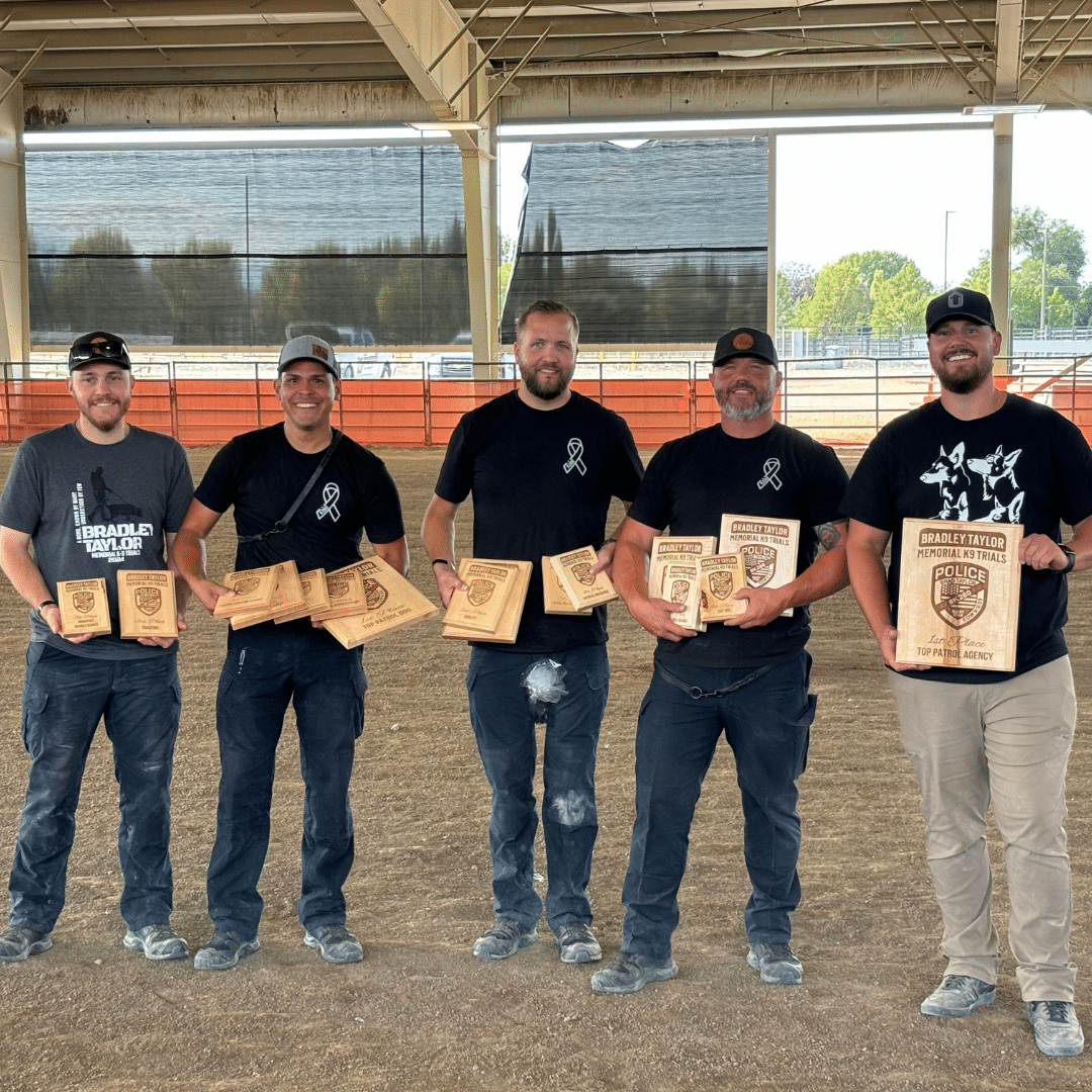 Five men standing in a row, each holding wooden awards. They are under a large open pavilion with a dirt floor. All are wearing casual clothing and hats, and they appear to be smiling.