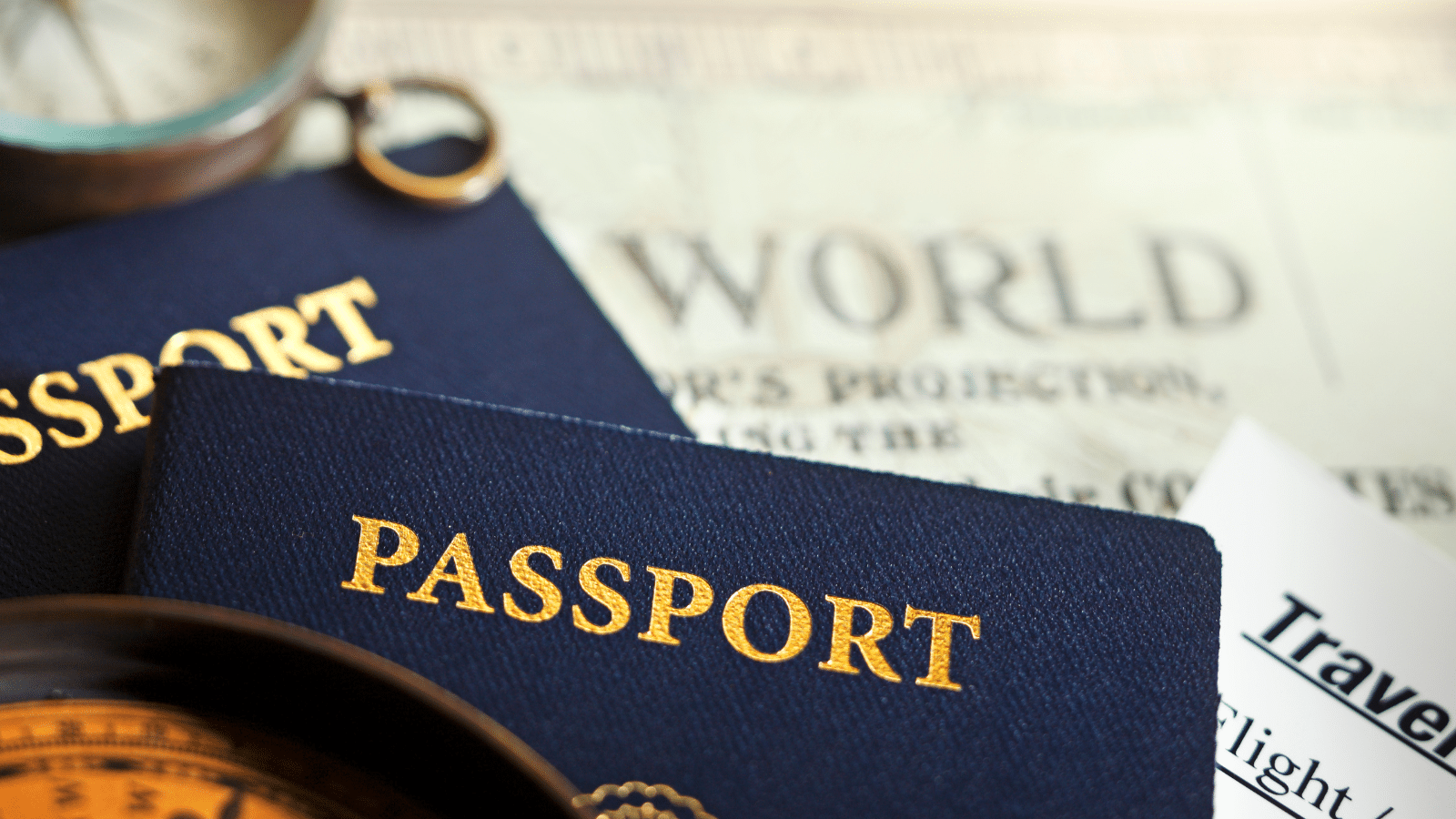 Close-up of two blue passports with gold lettering on top of a map with the word WORLD visible. A partial travel document and a compass are also in the frame, suggesting a theme of travel and exploration.