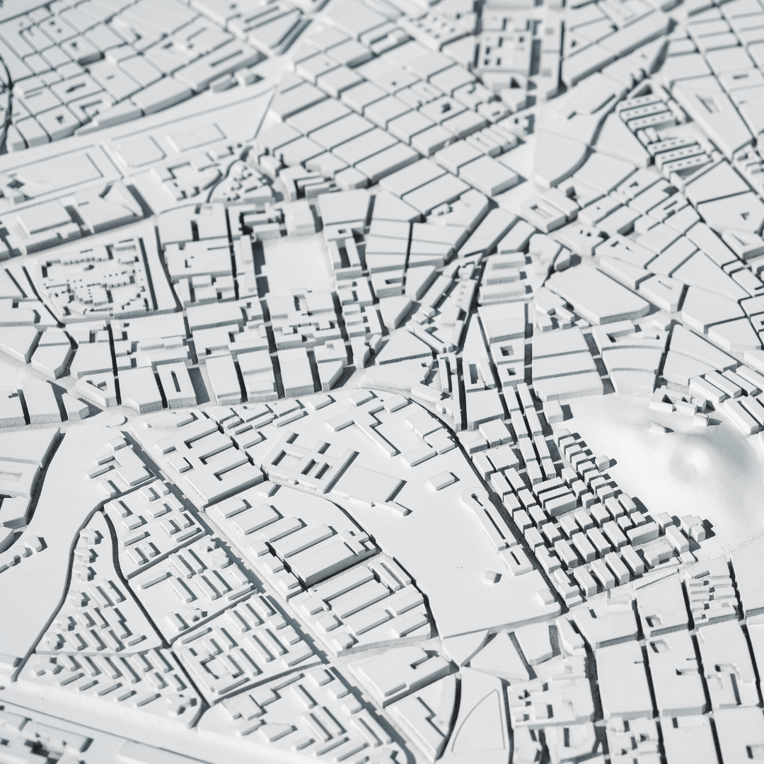 A detailed architectural model of an urban area, depicting various city blocks, streets, and buildings in a white, monochromatic style. The layout is intricate, showing both densely populated areas and more open spaces.