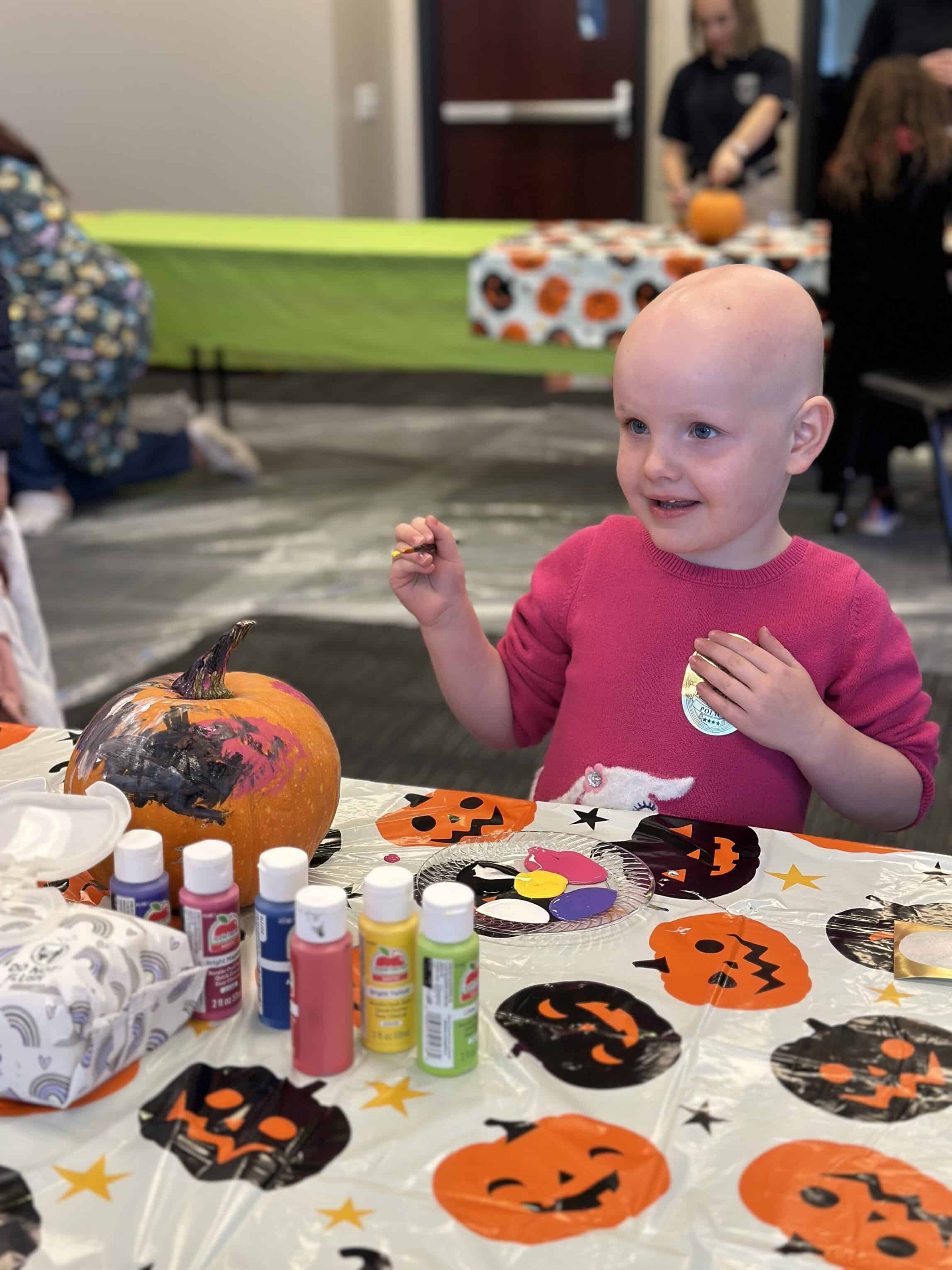 A child in a pink sweater paints a pumpkin at a festive table covered with a Halloween-themed tablecloth. Various paints and crafting materials are scattered around. In the background, other participants engage in similar activities.