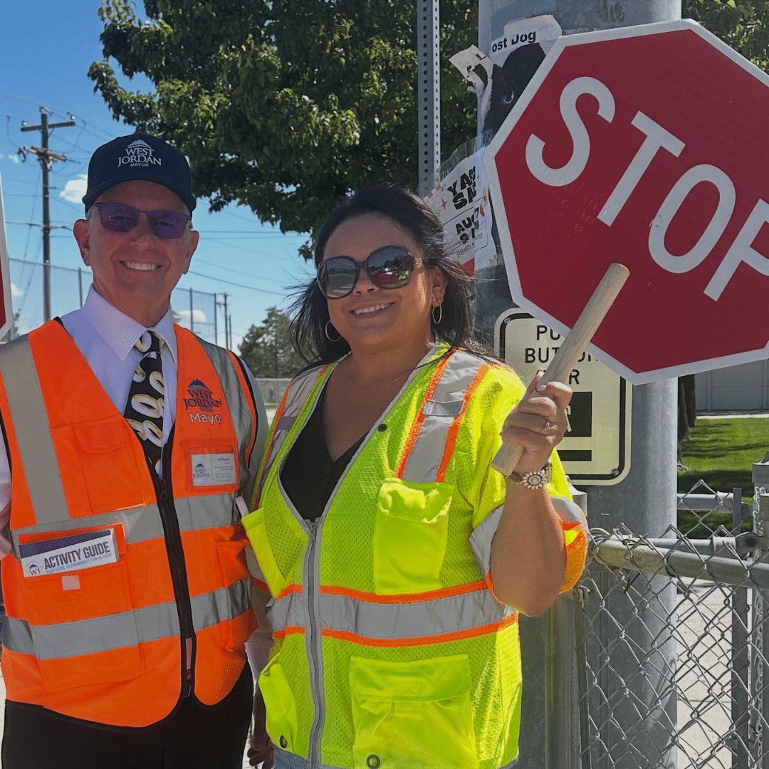 Two people wearing safety vests stand outdoors. One holds a small stop sign. They are both smiling, with shades and hats on under a sunny day. A fence, trees, and traffic signs are in the background.