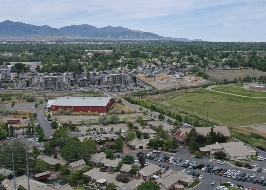 Aerial view of a suburban area with houses, apartment buildings, and a large red building. Roads intersect the neighborhood surrounded by green spaces, with mountains visible in the background under a cloudy sky.