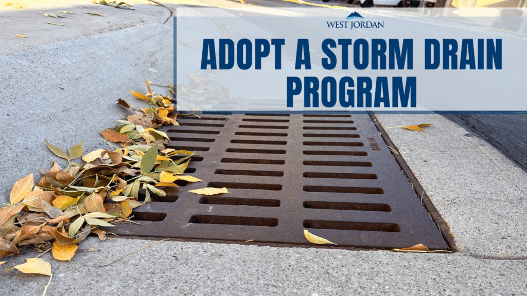 A metal storm drain on a street corner is partially covered with fallen leaves. A sign above reads West Jordan - Adopt a Storm Drain Program.