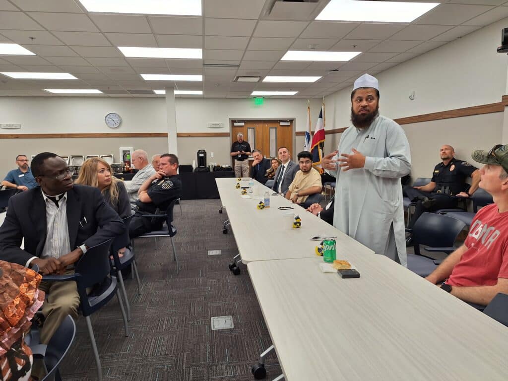 A man in a white robe speaks to a diverse group seated around a conference table. The room has a flag and a clock on the wall. People listen attentively, with some police officers present.