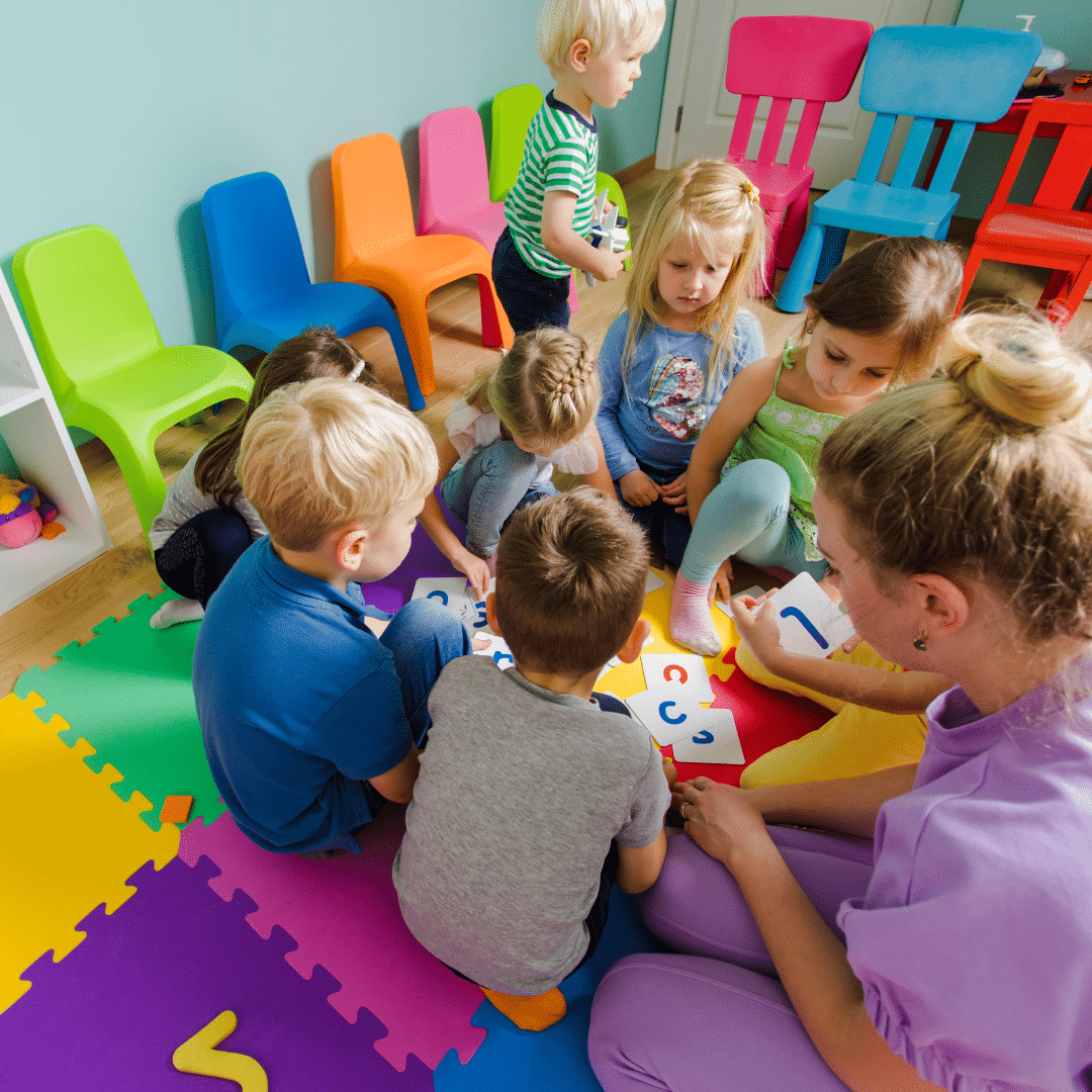 Children sitting in a circle on colorful foam mats, engaging in an educational activity with large flashcards containing letters and numbers. Brightly colored chairs line the walls in a cheerful classroom setting.