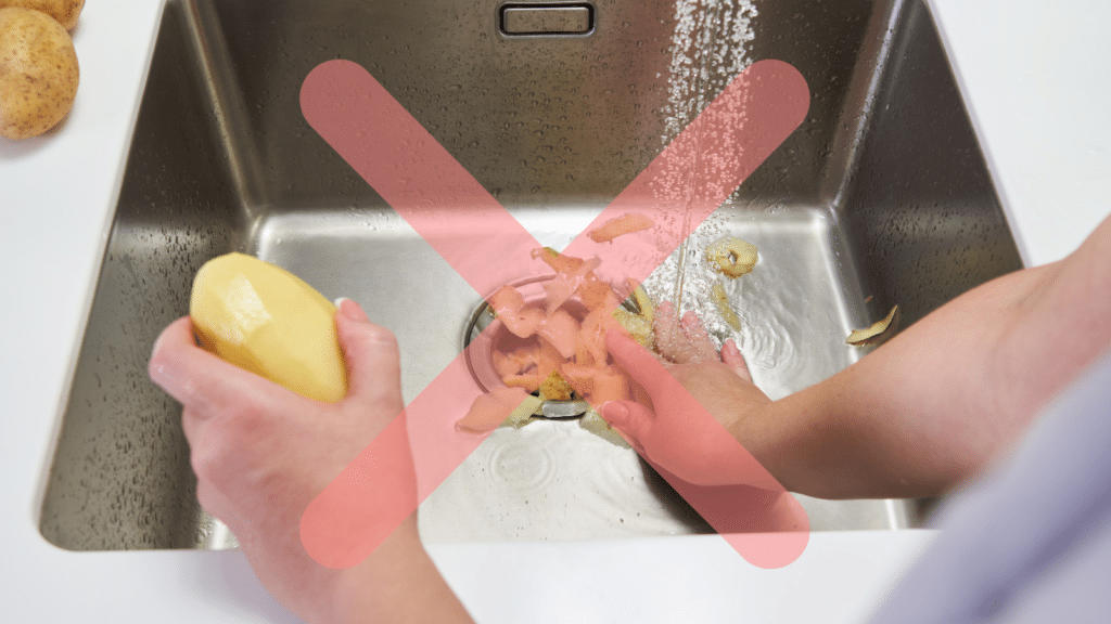 A person peels a potato over a sink with potato peels near the drain. A red X overlays the image, indicating not to dispose of peels in the sink.