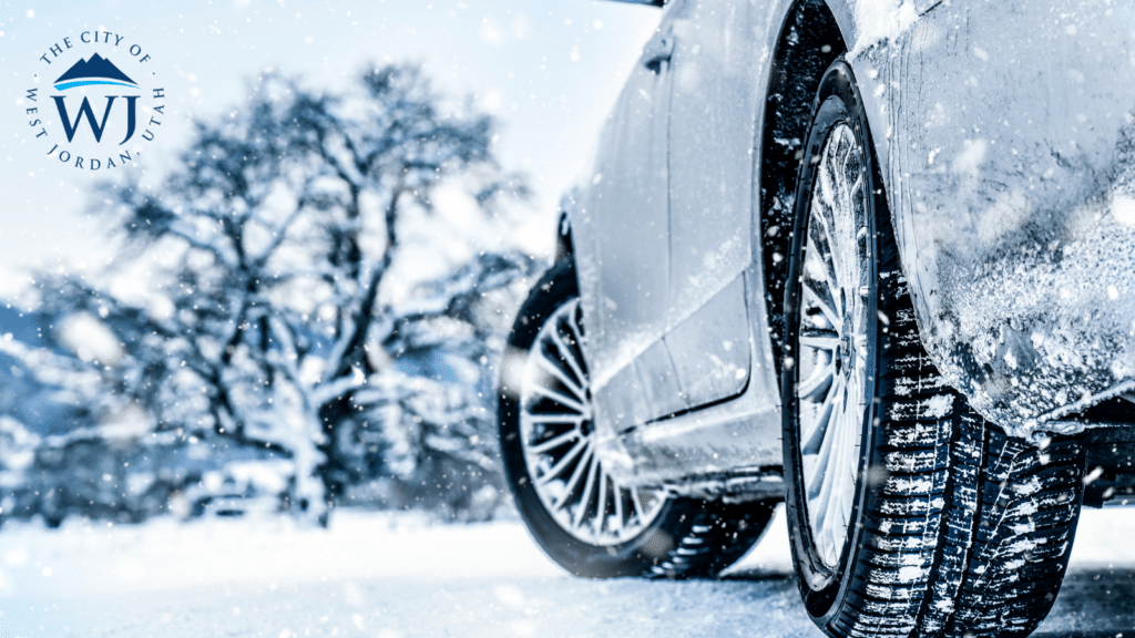 Close-up of a cars tires driving on a snowy road with snow falling. A blurred background shows snow-covered trees. The City of West Jordan, Utah logo is in the top left corner.