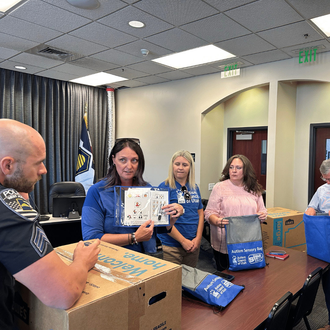 A group of people in a room, some wearing uniforms, stand around a table with boxes labeled Weighted Blanket. One woman holds a communication board, while others prepare sensory kits in blue bags for the Autism Sensory Initiative.