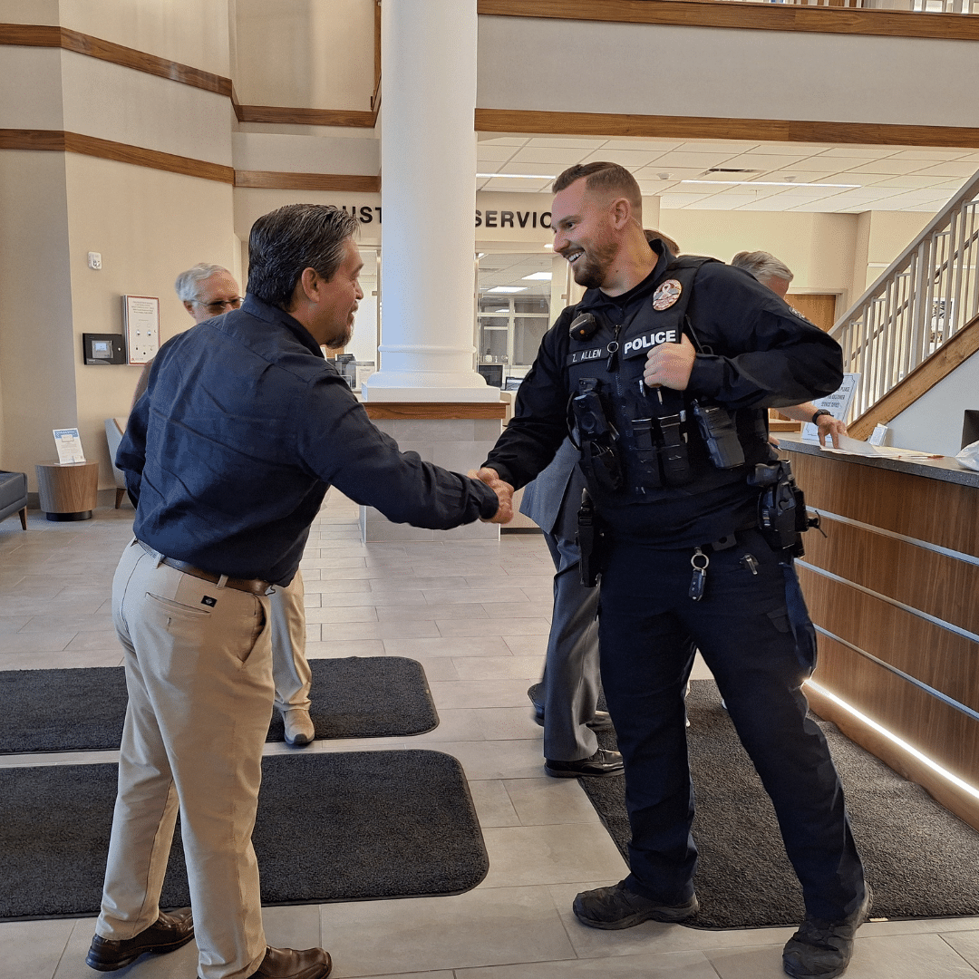 A man in a police uniform shakes hands with another man in a lobby. Two other people are in the background near a staircase. The setting appears to be a public or office space with tiled flooring.