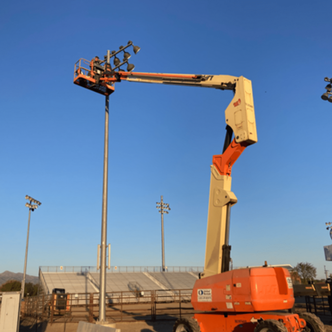 A person operates a cherry picker, working on stadium lights under a clear blue sky. The equipment is extended high above the bleachers in an outdoor sports field.