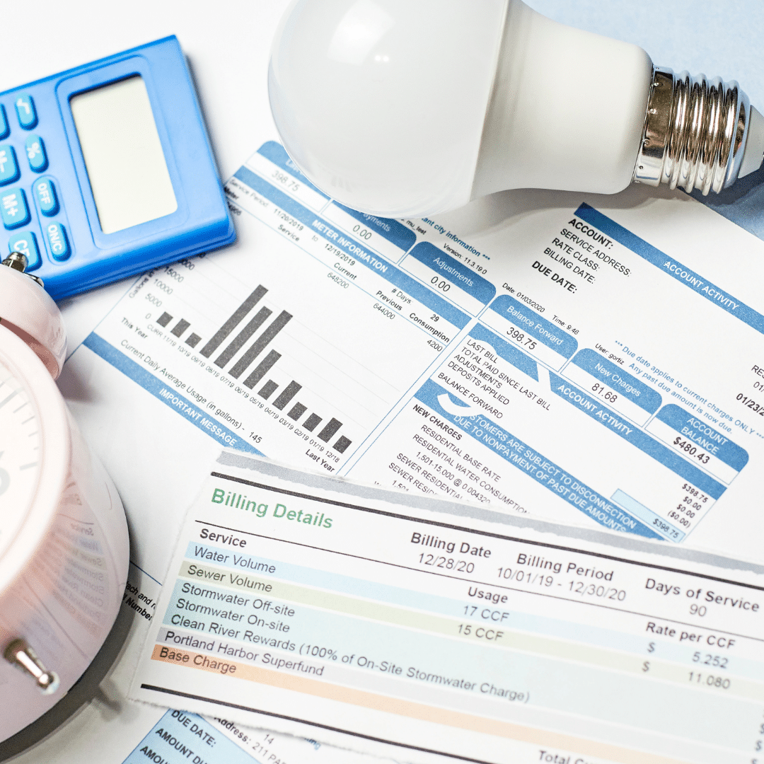 A cluttered desk with utility bills, a light bulb, a calculator, and a pink alarm clock. The bills feature graphs and tables with billing details. The scene suggests financial planning or utility cost assessment.