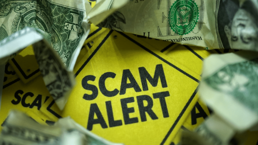 A yellow Scam Alert sign is surrounded by crumpled U.S. dollars, emphasizing caution about financial scams.