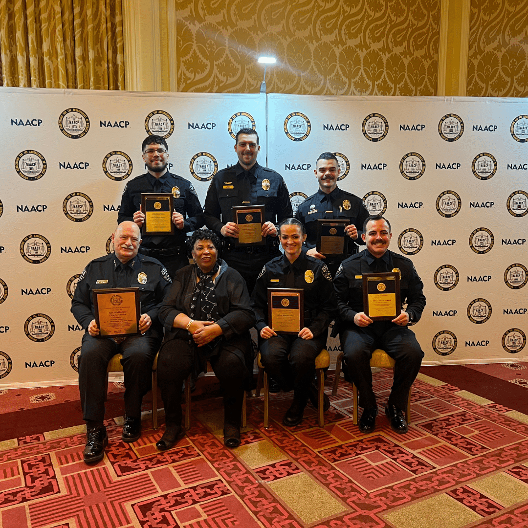 Seven people, six in police uniforms and one in business attire, pose with plaques in front of a backdrop featuring NAACP logos. They stand and sit on a patterned carpet in what appears to be a formal event setting.