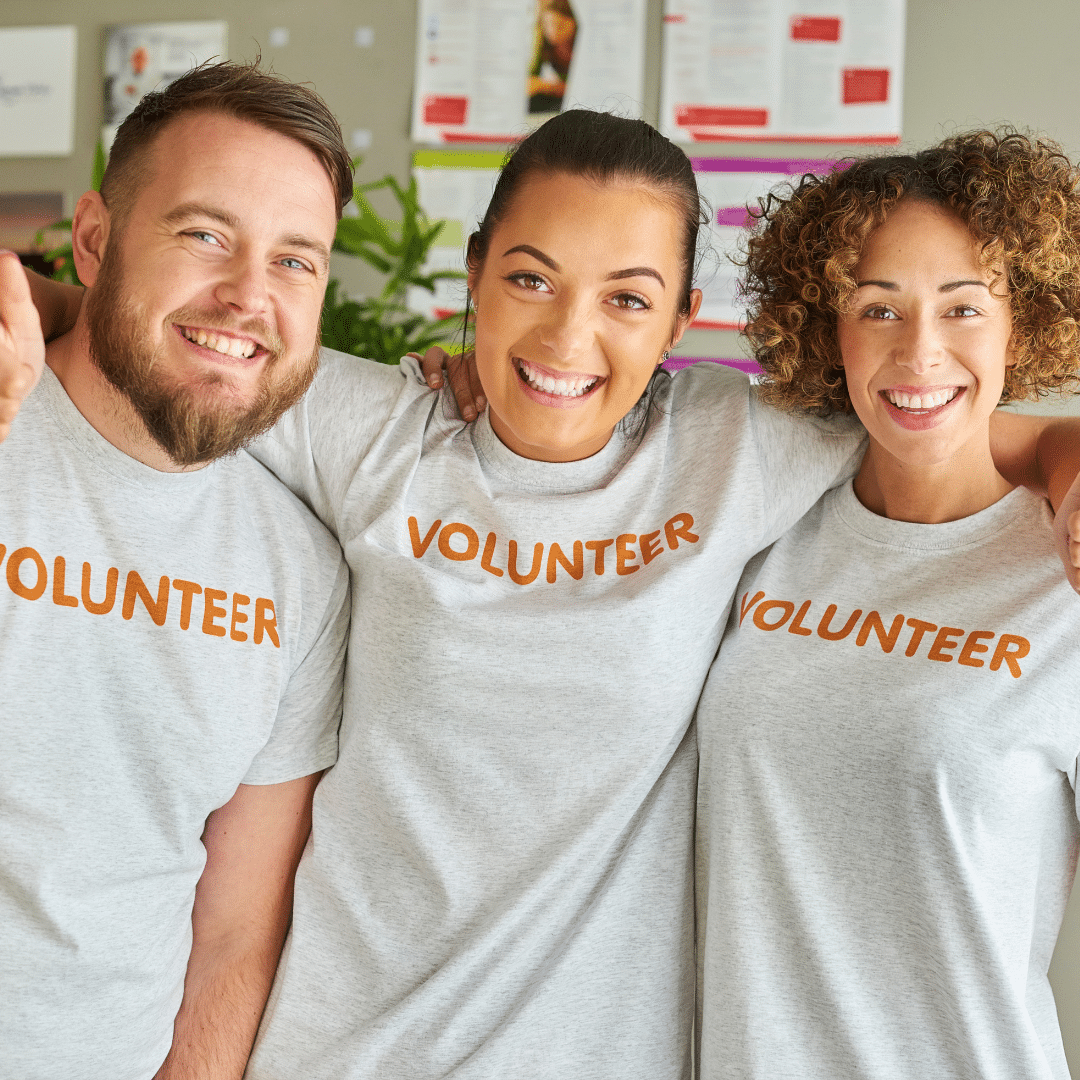 Three smiling individuals with arms around each other wearing gray VOLUNTEER T-shirts, stand indoors. They exude friendliness and teamwork in a cozy setting with colorful posters in the background.