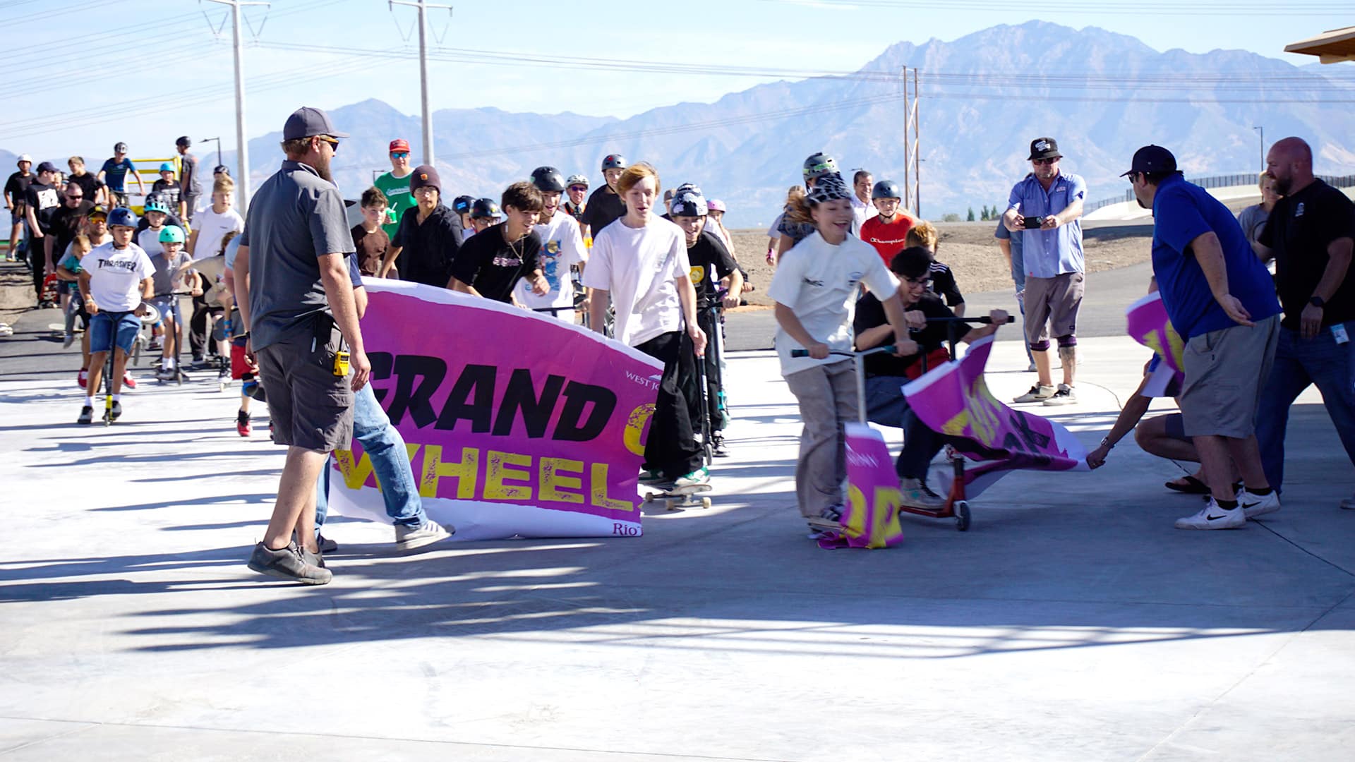 A group of people, including children on scooters and skateboards, crowd around banners during an outdoor event. They are on a concrete surface with mountains visible in the background. The atmosphere is lively and energetic.