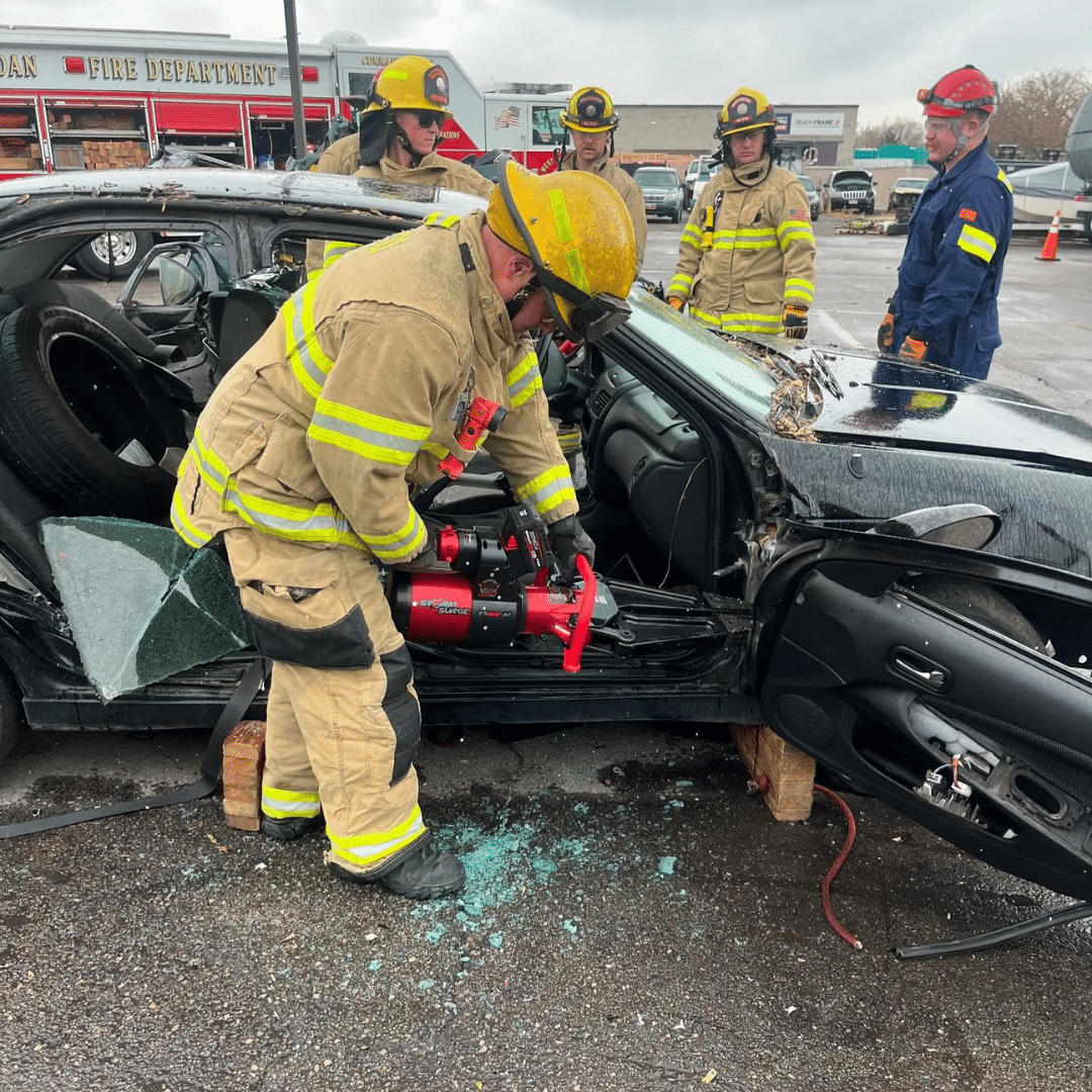 Firefighters in yellow protective gear use hydraulic rescue tools to open a black cars door in a parking lot. The car is stabilized on wooden blocks, and shattered glass is visible on the ground. Emergency vehicles are in the background.