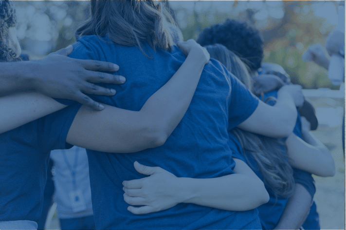 A group of people in blue shirts embracing in a supportive group hug outdoors. They have their arms around each other, creating a sense of community and connection. The background is blurred, suggesting a natural setting.