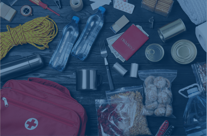 A flat lay of emergency preparedness items, including a red first aid kit, water bottles, canned food, a flashlight, snacks, a passport, a map, a rope, and utensils, all arranged on a dark wooden surface.