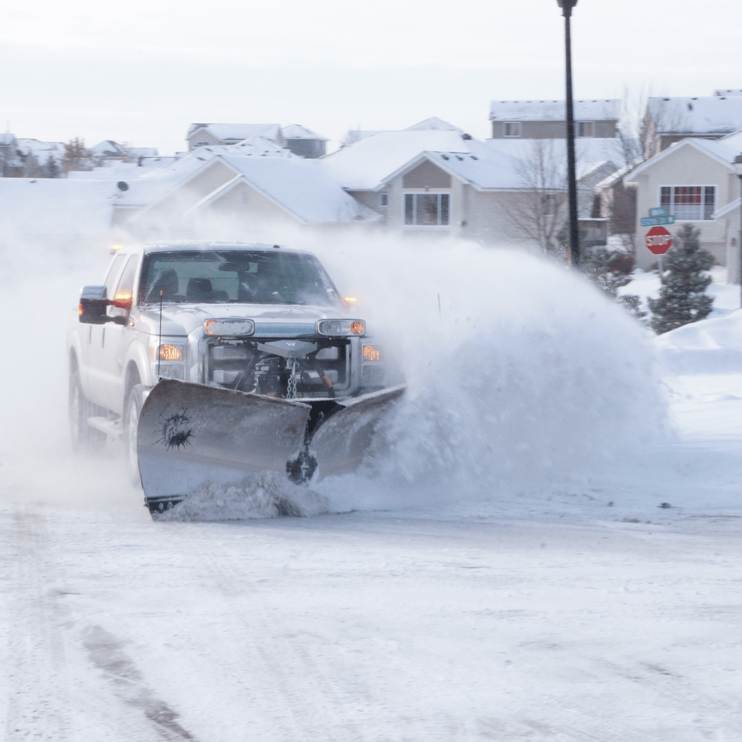 A large truck equipped with a snowplow is clearing snow on a residential street. Snow is billowing to the side as the plow moves forward. Houses and a stop sign are visible in the background under a cloudy sky.