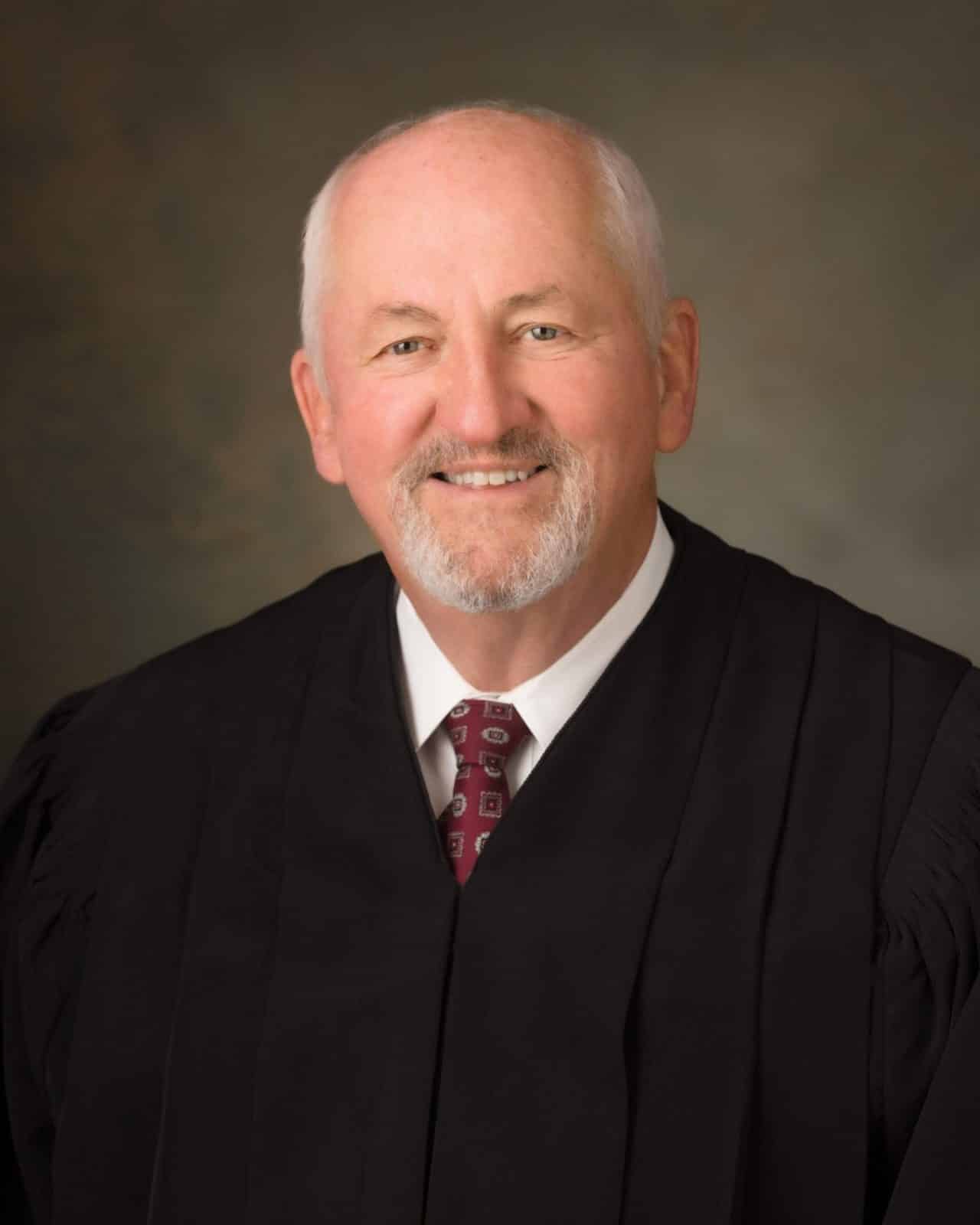 A smiling elderly man in a black judges robe and a patterned maroon tie poses against a neutral backdrop. He has short white hair, a mustache, and a goatee.