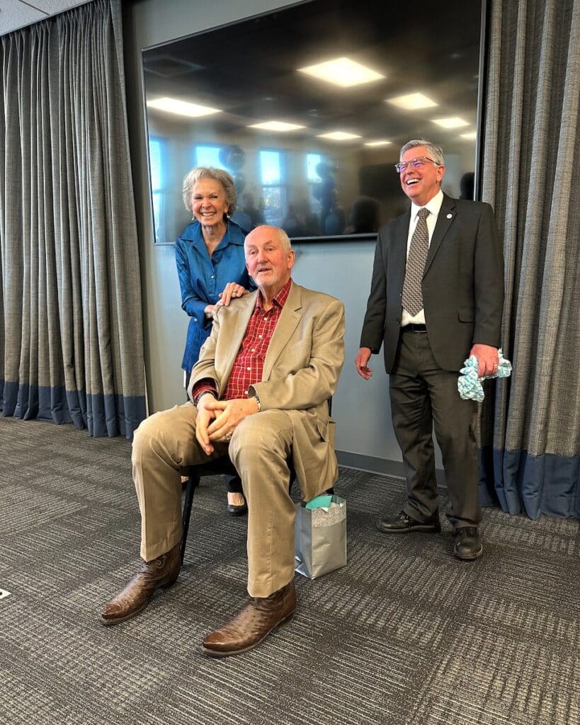 An older man sitting on a chair is smiling and wearing a beige suit with a red plaid shirt. A woman stands behind him with her arm on his shoulder, smiling. Another man in a suit stands beside them, also smiling. A large TV is on the wall behind them.