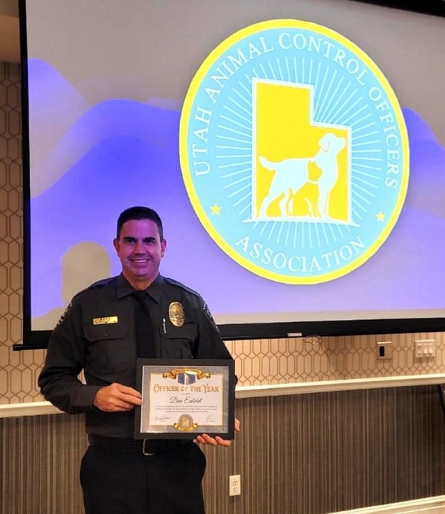 A uniformed man stands smiling, holding a framed Officer of the Year certificate. Behind him is a screen with the logo of the Utah Animal Control Officers Association, featuring a goat silhouette and a map of Utah.