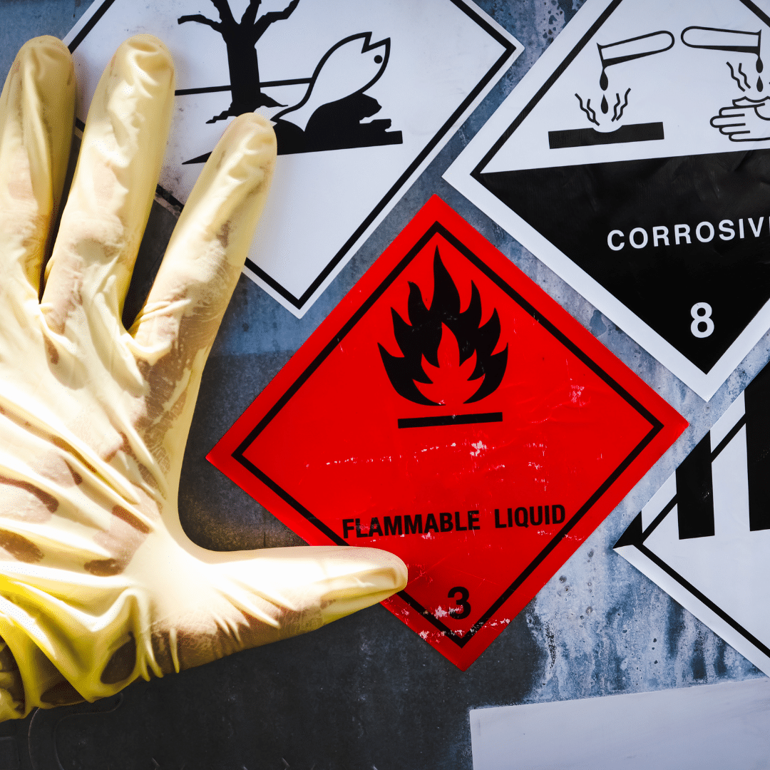 A gloved hand points at a red diamond-shaped hazard sign with a flame icon, labeled Flammable Liquid 3. Other labels nearby include symbols for corrosive substances.