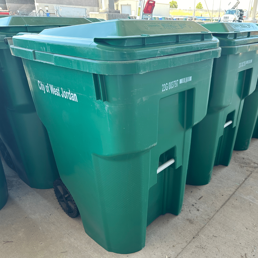 A row of green wheeled garbage bins stands on a concrete surface. The bins are labeled City of West Jordan with a serial number. Sunlight casts shadows from nearby structures.