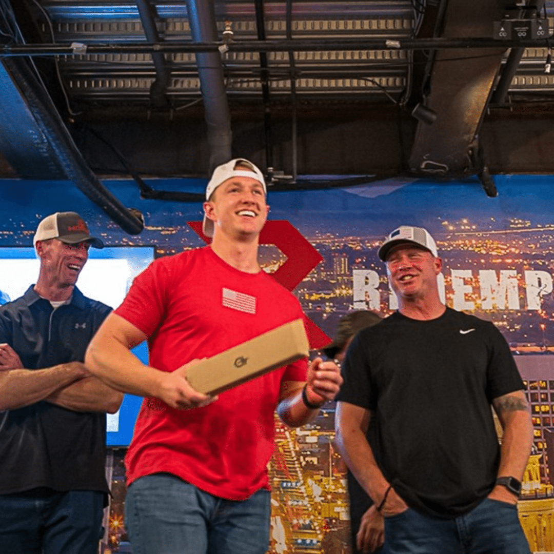 Three people are standing indoors, smiling and laughing. One person in the center holds a box. They are in casual clothes and wearing caps. The background shows a large sign with city imagery.