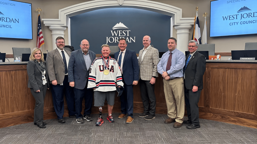 A group of eight people, including a man wearing a USA hockey jersey and gold medals, stand together in front of a West Jordan City Council sign. They are in a formal setting with flags and a wood-paneled desk behind them.