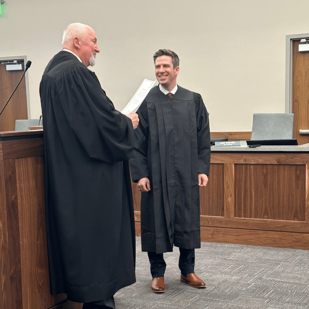 Two men in black robes stand facing each other in a courtroom. The man on the left holds a document and is speaking, while the man on the right smiles. Wood-paneled desks and chairs are in the background.