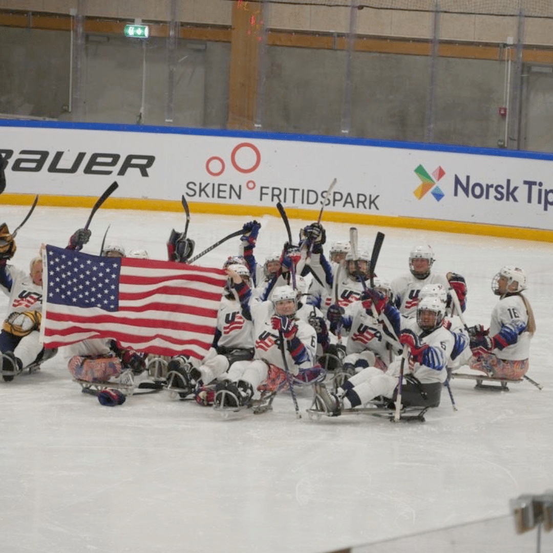 A group of sled hockey players on the ice, wearing white jerseys with red and blue details. They are holding hockey sticks up and displaying a large American flag. The backdrop includes advertisements and a wall with windows.