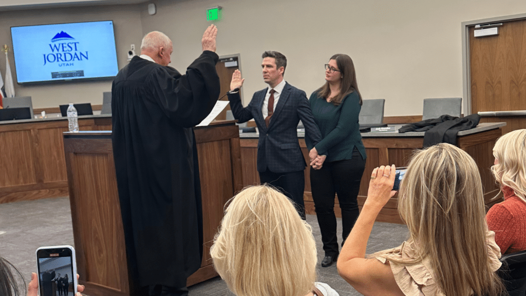 A judge administers an oath to a man in a suit while a woman stands beside him in a courtroom setting. The audience watches, some taking photos. A screen displays the text West Jordan Utah.