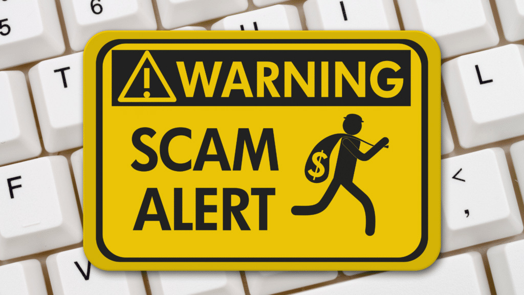 A yellow sign with a triangular warning symbol and bold text reading WARNING SCAM ALERT is set against a backdrop of white computer keyboard keys. A silhouette of a person carrying a bag with a dollar sign is depicted.