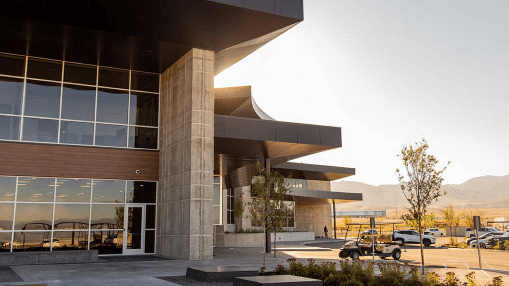 Modern building exterior with large windows and concrete pillars. Sunlight filters from behind the structure. Landscaped gardens and a small utility vehicle are visible in front. Mountains appear in the background under a clear sky.