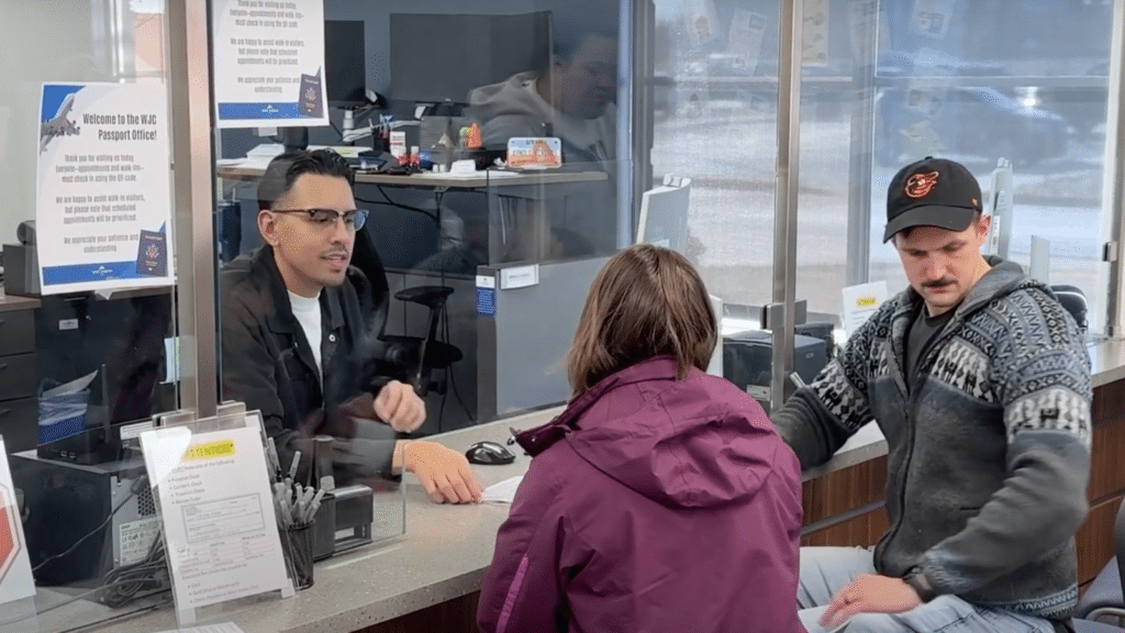 Two people sit at a service counter speaking with an employee through clear glass. The man behind the counter is explaining something. Visible signs and a computer are on the desk. Both customers wear casual clothing, including a sweater and a cap.