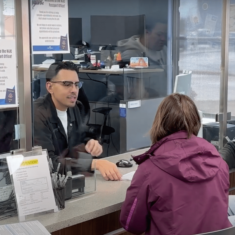 A person in a purple jacket sits at a counter speaking with a worker behind a glass barrier in an office setting. The worker is seated, wearing glasses and a dark jacket. Papers and office supplies are visible on the counter.